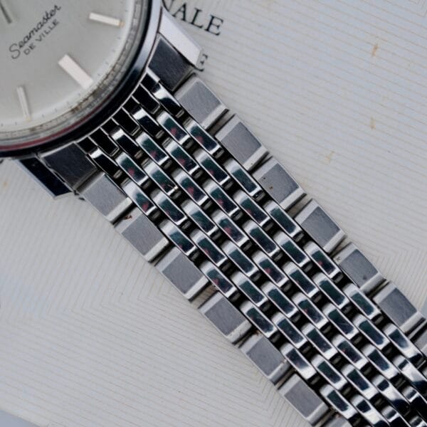 Close-up of a silver metal watch band attached to a round 1968 Omega Seamaster Deville 165.020 watch face, displaying part of the text Seamaster De Ville on a light textured background.