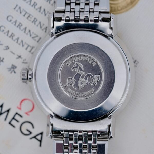A close-up of the back of a 1968 Omega Seamaster Deville 165.020 wristwatch, showing the engraved Seamaster Waterproof seahorse logo and metal bracelet, with part of an Omega document in the background.