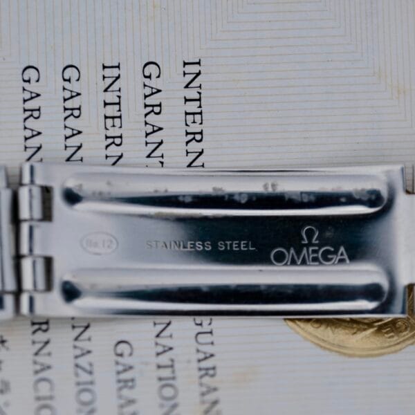 A close-up of the 1968 Omega Seamaster Deville 165.020’s stainless steel clasp highlights the engraved Omega logo, STAINLESS STEEL, No.12, with part of a printed warranty card visible underneath.