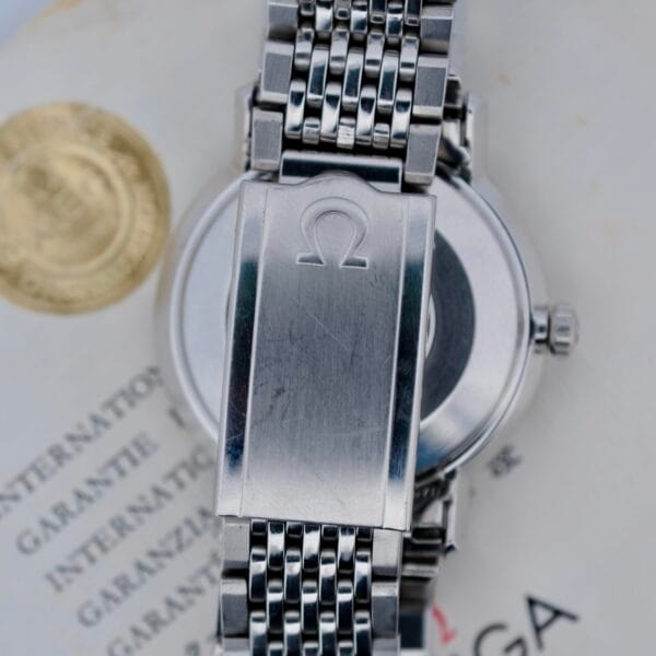 A close-up of the back of a stainless steel 1968 Omega Seamaster Deville 165.020 with a metal bracelet, showing the engraved Omega logo on the clasp. Part of a warranty certificate with a gold seal is visible underneath.