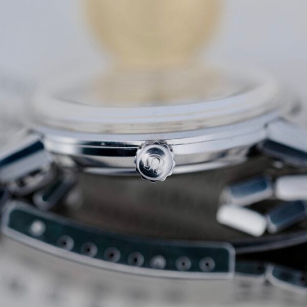 Close-up of the side crown on the 1968 Omega Seamaster Deville 165.020 wristwatch, highlighting its detailed logo. The metallic case is in focus, with part of the strap blurred in the background.