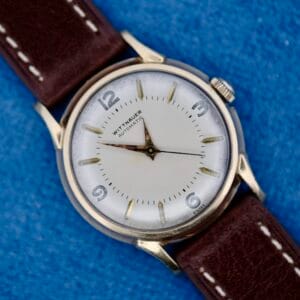 A 1950s Wittnauer Automatic Teardrop Lugs wristwatch with a white dial, silver case and hour markers, and brown leather strap is displayed on a blue textured surface, showing the time at approximately 9:14:04.