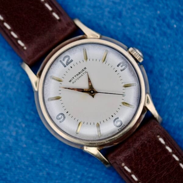 A 1950s Wittnauer Automatic Teardrop Lugs wristwatch with a white dial, silver case and hour markers, and brown leather strap is displayed on a blue textured surface, showing the time at approximately 9:14:04.