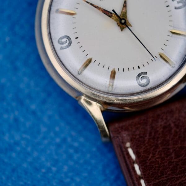 A close-up of the 1950s Wittnauer Automatic Teardrop Lugs wristwatch with a white dial, gold hands, black numerals (showing 9, 6, and 3), a brown leather strap, and a blue textured background.