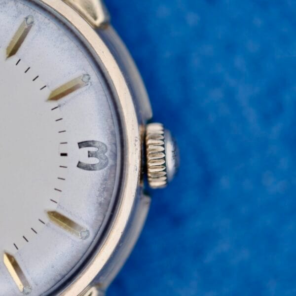 Close-up of the 1950s Wittnauer Automatic Teardrop Lugs watch, highlighting its crown and raised hour markers at 3 o’clock against a blue textured background. Minute markers adorn the dial for precise timekeeping.