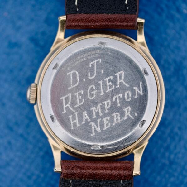 A close-up of the back of a 1950s Wittnauer Automatic Teardrop Lugs wristwatch with a metal case engraved D.J. Regier Hampton Nebr. It features a black and brown leather strap and is set against a blue background.