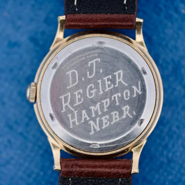 A close-up of the back of a 1950s Wittnauer Automatic Teardrop Lugs wristwatch with a metal case engraved D.J. Regier Hampton Nebr. It features a black and brown leather strap and is set against a blue background.