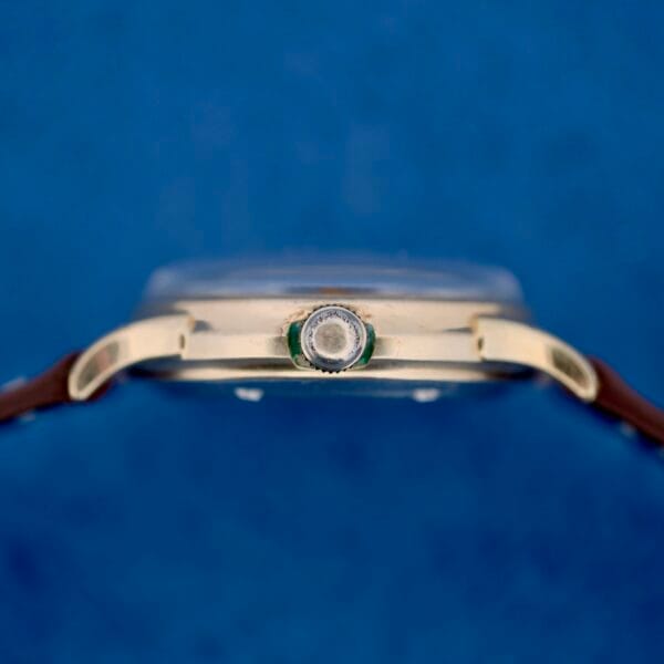 Close-up side view of a 1950s Wittnauer Automatic Teardrop Lugs wristwatch with a brown leather strap, gold-colored case, and center crown against a blue background. The watch face is not visible in this image.