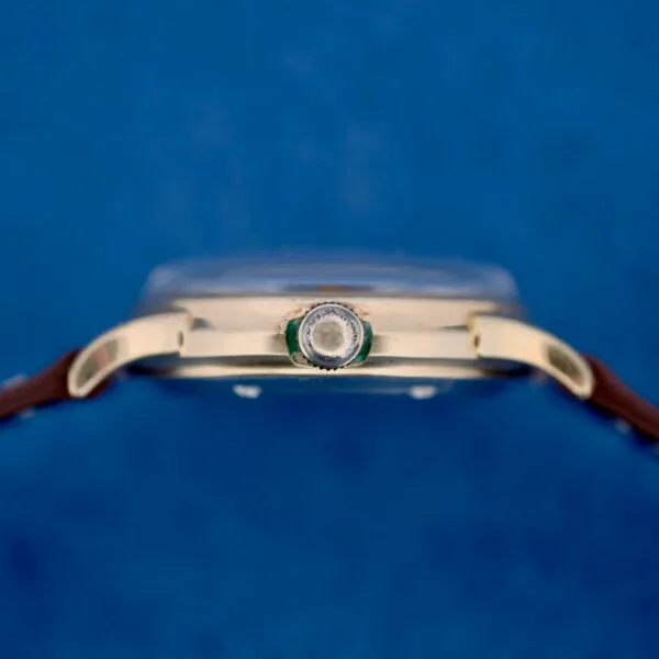 Close-up side view of a 1950s Wittnauer Automatic Teardrop Lugs wristwatch with a brown leather strap, gold-colored case, and center crown against a blue background. The watch face is not visible in this image.