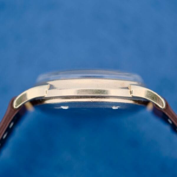 Side view of the 1950s Wittnauer Automatic Teardrop Lugs wristwatch with a gold case, dome-shaped crystal, and brown leather strap against a blue background.