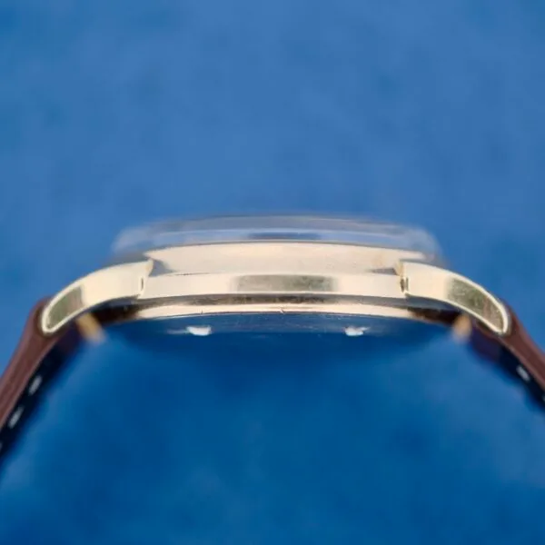 Side view of the 1950s Wittnauer Automatic Teardrop Lugs wristwatch with a gold case, dome-shaped crystal, and brown leather strap against a blue background.