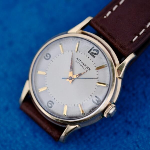 A close-up of a 1950s Wittnauer Automatic Teardrop Lugs wristwatch with a white dial, gold hour markers, and a brown leather strap, set against a blue textured background. The time displayed is 4:08:38.