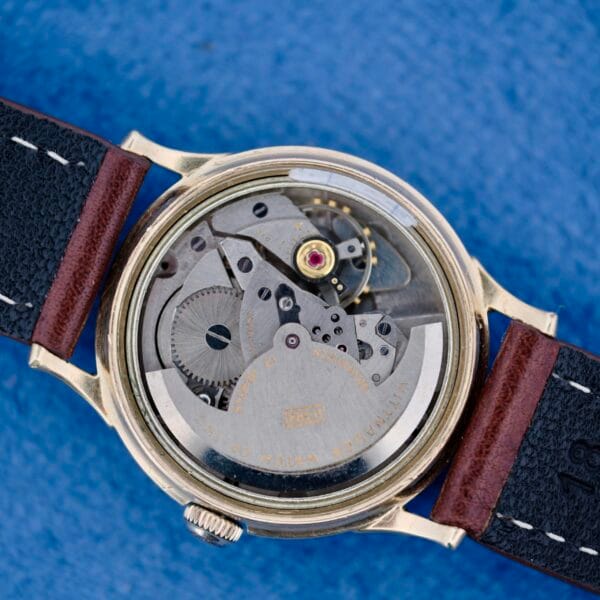 Close-up of the back of a 1950s Wittnauer Automatic Teardrop Lugs with the case removed, revealing gears and inner workings. The watch features a black leather strap with brown ends, white stitching, and rests on a blue surface.