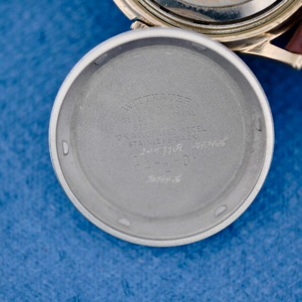The inside case back of a 1950s Wittnauer Automatic Teardrop Lugs wristwatch is shown, featuring engraved serial numbers and specifications, resting on a blue textured surface.