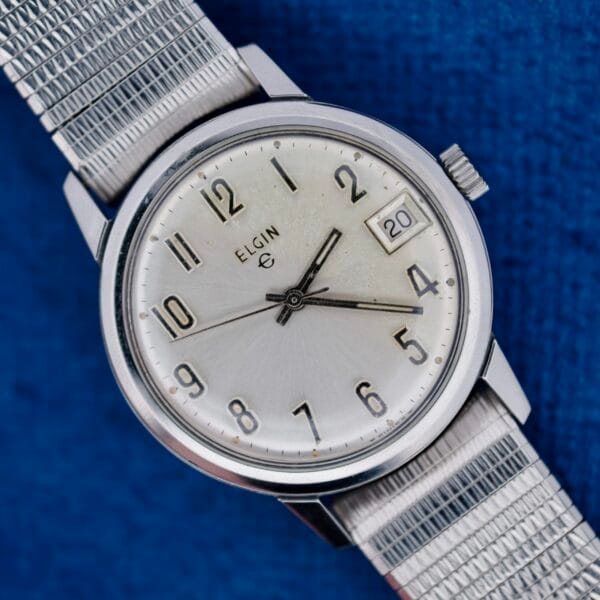 An Elgin Date Stainless Steel Arabic Numerals watch with a white face, black numerals, date window showing 20, and metal mesh band is shown on a blue textured surface. The time reads about 7:08:42.