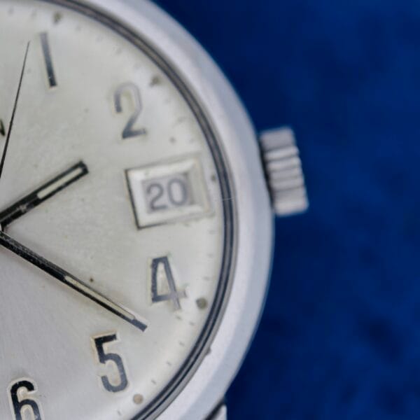 Close-up of an Elgin Date Stainless Steel Arabic Numerals wristwatch showing 8:23:19 and date 20, set against a blue background.