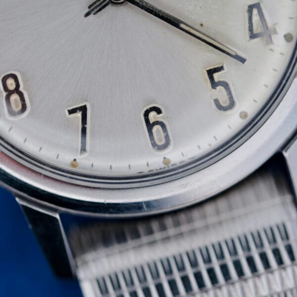 Close-up of the Elgin Date Stainless Steel Arabic Numerals watch, highlighting its silver analog face showing numbers 6, 7, and 8 with black hands near 4:30. The metal mesh band and part of the watch face are in sharp focus.