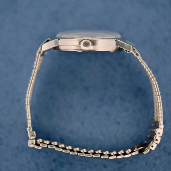 The Elgin Date Stainless Steel Arabic Numerals wristwatch is displayed on its side against a blue background, highlighting the crown and the side profile of the stainless steel case and bracelet links.