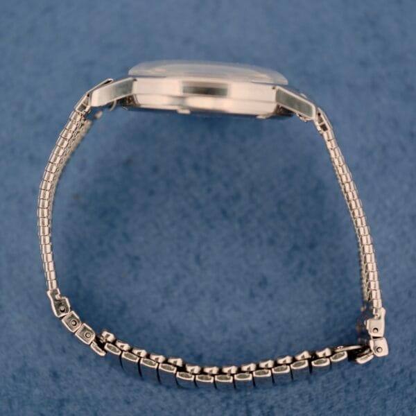 The Elgin Date Stainless Steel Arabic Numerals watch is displayed in profile against a blue background, featuring a round, slightly raised case and a partially open metal link band forming a semicircle.