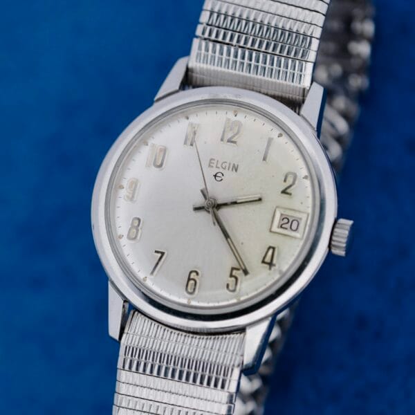 A close-up of the Elgin Date Stainless Steel Arabic Numerals watch, featuring a silver metal band, silver dial, Arabic numerals, and a date window showing 20, set against a blue background.