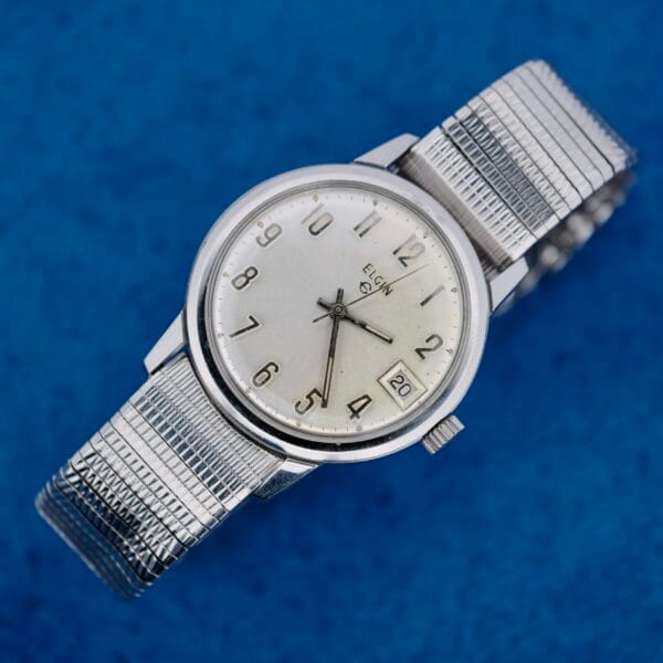 An Elgin Date Stainless Steel Arabic Numerals wristwatch with a silver dial, date at 3 oclock, and gold hour numbers is shown against a blue background, displaying the time 8:18:26.