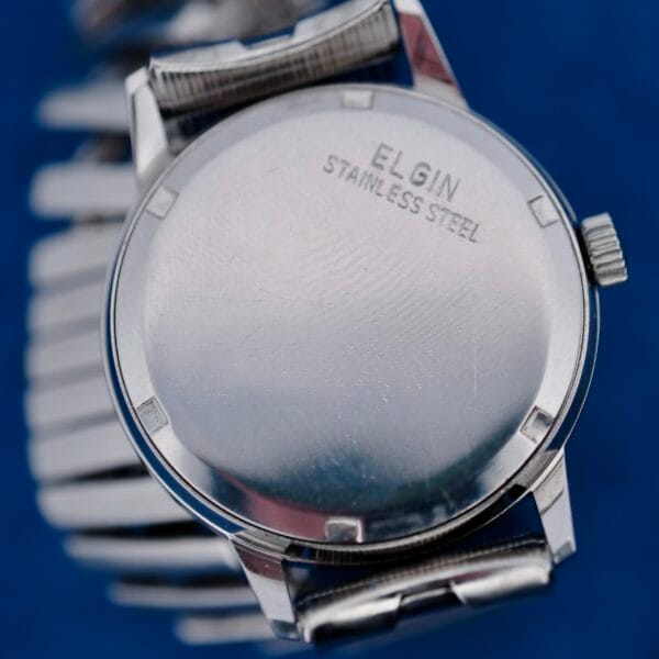 Close-up of the Elgin Date Stainless Steel Arabic Numerals watch with a metal band, showing the engraved back against a blue background.