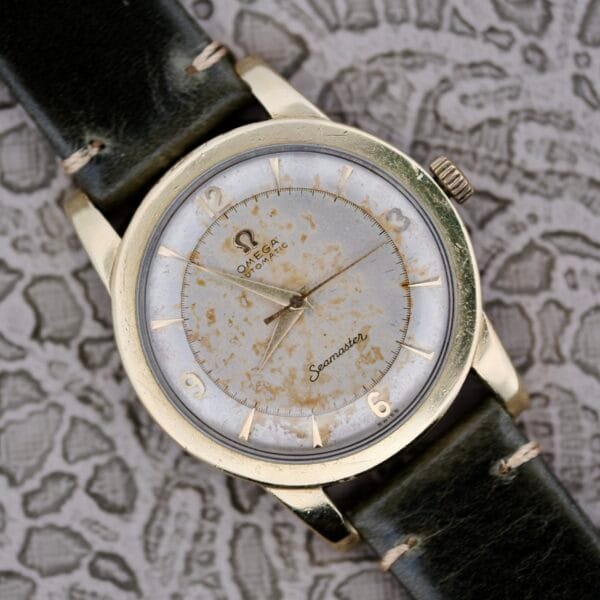 An Omega Seamaster Automatic 2577-17 with a tropic dial, gold-tone case, aged white and gold dial with Arabic numerals, and black leather strap is displayed on a patterned surface.
