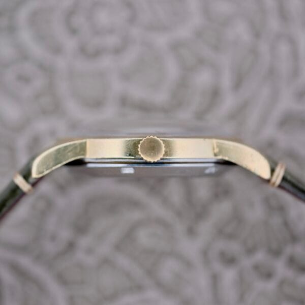 Side view of the Omega Seamaster Automatic 2577-17 Tropic Dial wristwatch with a gold-colored crown, leather strap, resting on a patterned gray surface.