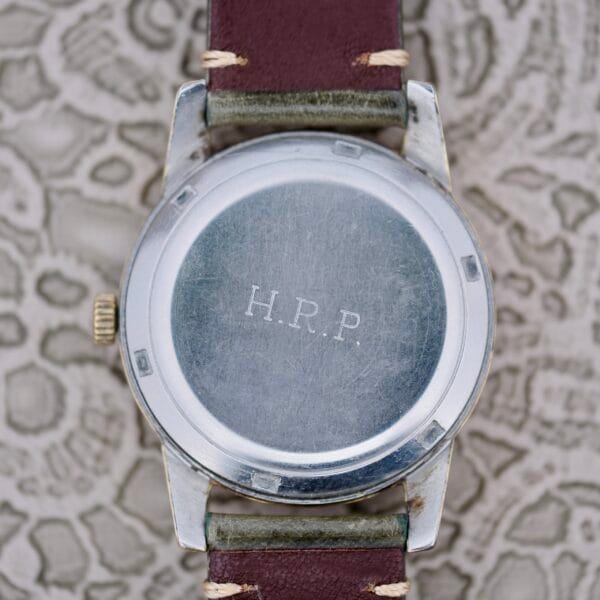 A close-up of the back of an Omega Seamaster Automatic 2577-17 Tropic Dial wristwatch with a brown leather strap. The metal case is engraved “H.R.P.” and rests on patterned neutral-toned fabric.