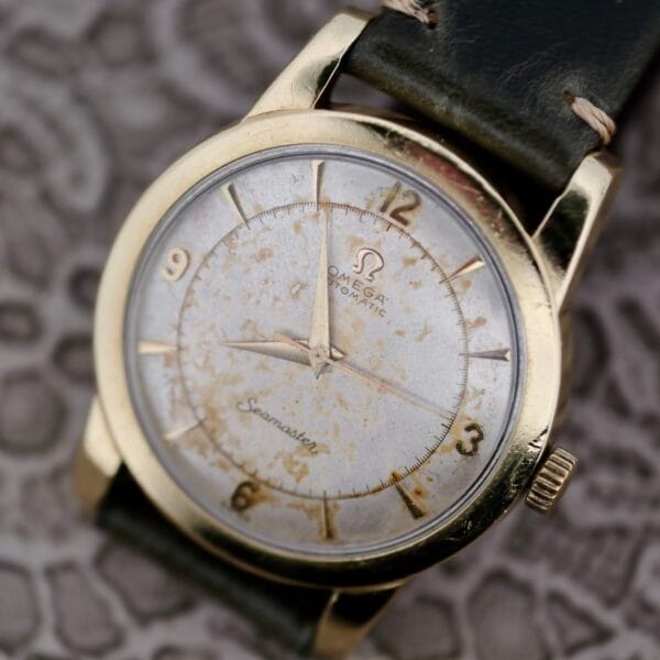An Omega Seamaster Automatic 2577-17 Tropic Dial with a worn gold case, aged white dial showing visible patina, Arabic numerals at 12, 3, 6, and 9, and a dark leather strap rests on a patterned surface.