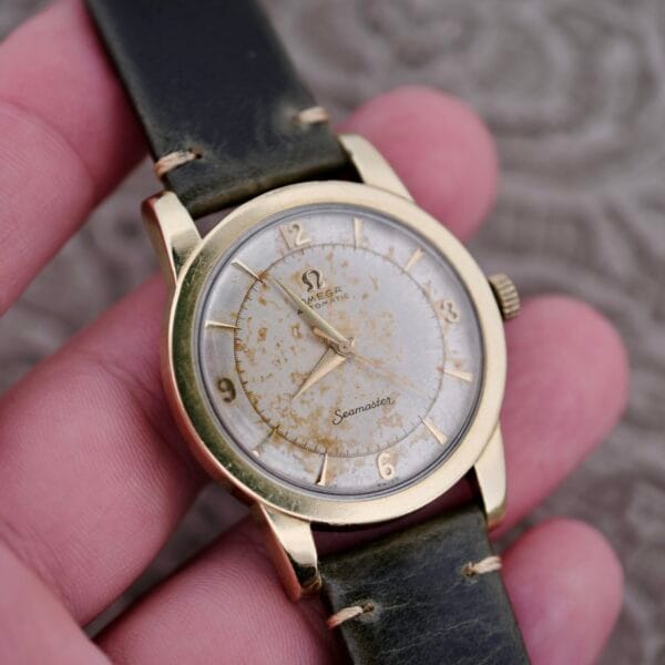A hand holds an Omega Seamaster Automatic 2577-17 Tropic Dial with a gold case, aged white dial, gold hour markers, and a dark green leather strap.