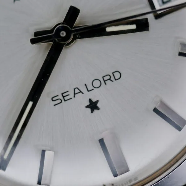 Close-up of the Benrus Automatic Sea Lord Date watch face, featuring black hands, rectangular hour markers, SEA LORD text above a small star, all set against a silver brushed metal background.