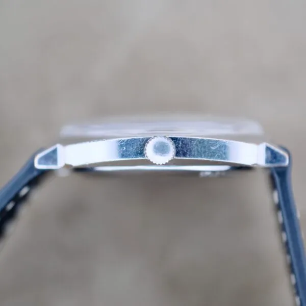 Close-up side view of a Benrus Automatic Sea Lord Date wristwatch with a round metal crown, visible case scratches, and black leather straps, set against a blurred beige background.