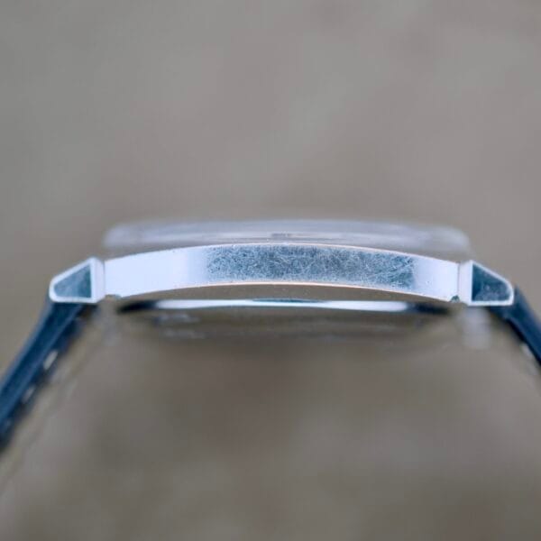 Side view of a Benrus Automatic Sea Lord Date case showing visible scratches on the metal, paired with a dark strap against a blurred beige background.