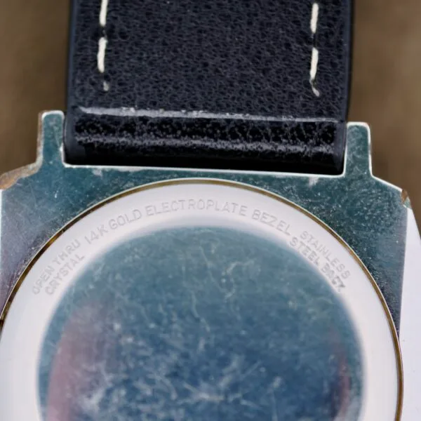 Close-up of the back of a Benrus Automatic Sea Lord Date wristwatch showing text: OPEN THRU CRYSTAL 14K GOLD ELECTROPLATE BEZEL STAINLESS STEEL BACK. The watch features a black leather strap with white stitching.
