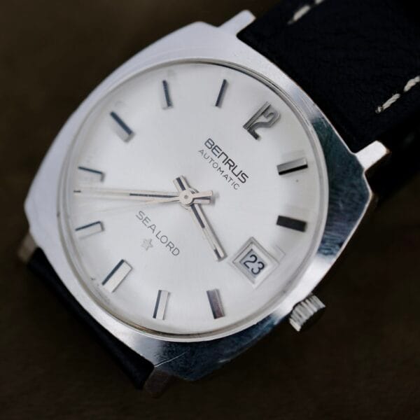 A close-up of a Benrus Automatic Sea Lord Date wristwatch with a silver dial, black hour markers, date at 23, and black leather strap. The time shown is about 8:42:36.