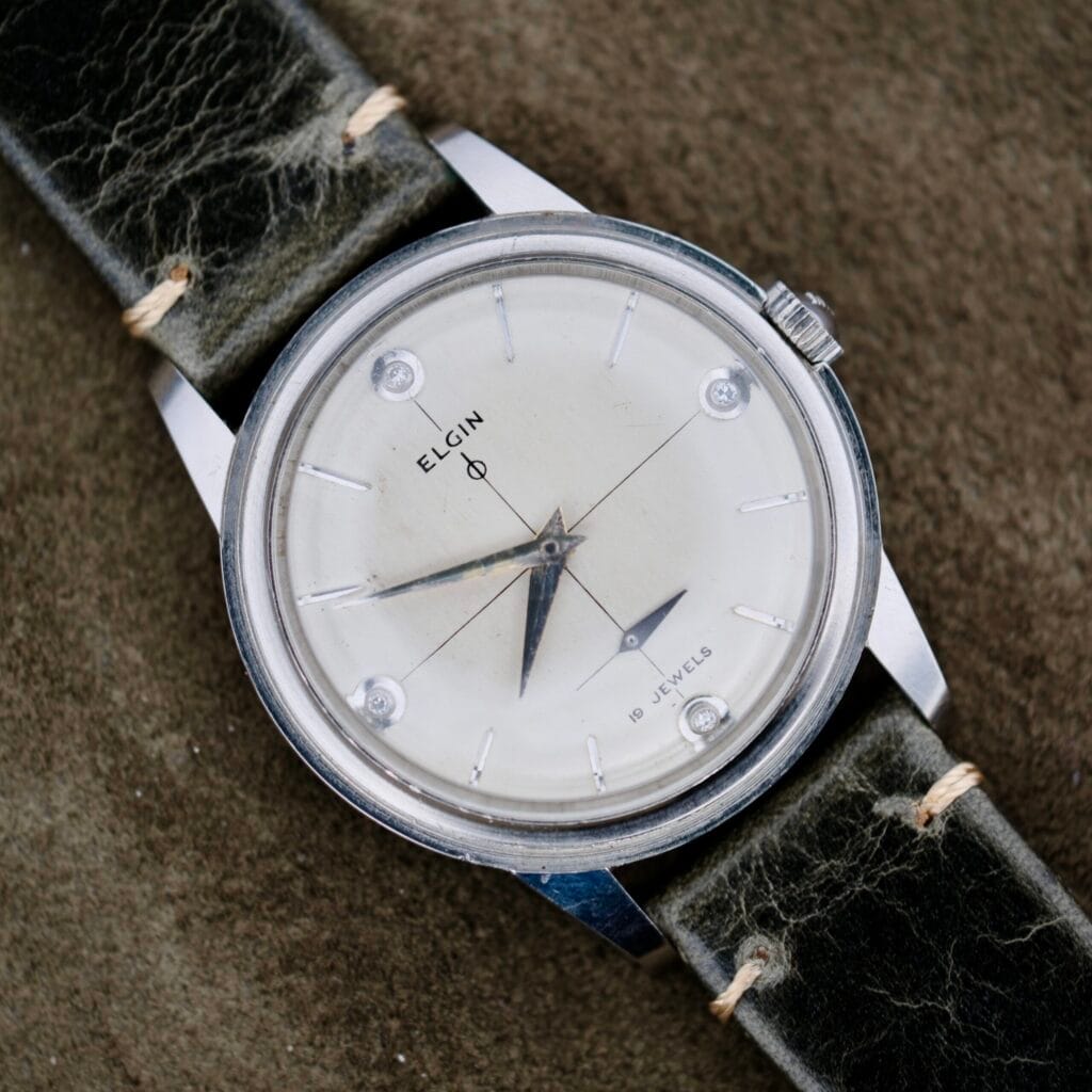 The Elgin 19 Jewels Manual Wind Diamond Indicies wristwatch features a white dial, silver case, worn black leather strap, and is powered by a precise 19 jewels manual wind movement.