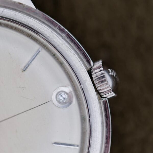 Close-up of the Elgin 19 Jewels Manual Wind Diamond Indicies wristwatch, featuring a silver face with minimalist hour markers, a diamond accent near the edge, and a ridged crown on the right. Background is blurred.