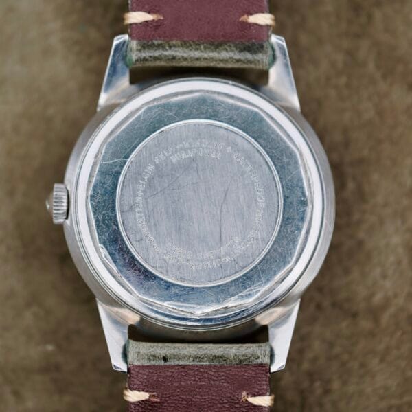 A close-up of the Elgin 19 Jewels Manual Wind Diamond Indicies watch, showing its worn silver back, green fabric strap with brown leather ends and stitching, resting on an olive-green textured surface.