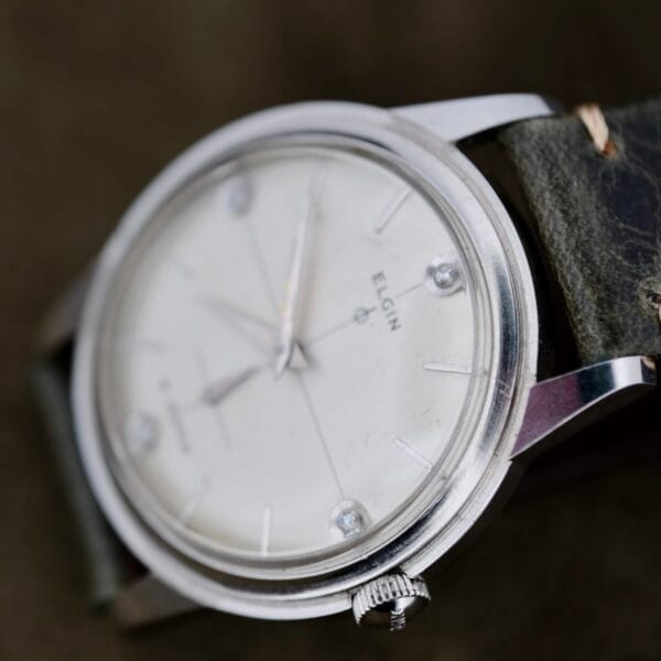 A close-up of an Elgin 19 Jewels Manual Wind Diamond Indicies wristwatch with a white face, silver hour markers, and a black leather strap. The watch is slightly angled, displaying the Elgin brand name on the dial.