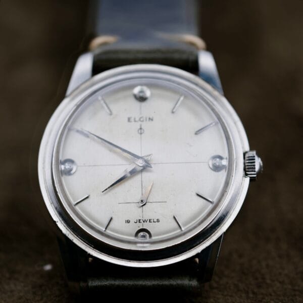 A close-up of the Elgin 19 Jewels Manual Wind Diamond Indicies watch, featuring a white dial, silver markers, two main hands, a small subdial, 19 JEWELS at the bottom, and a dark leather strap.