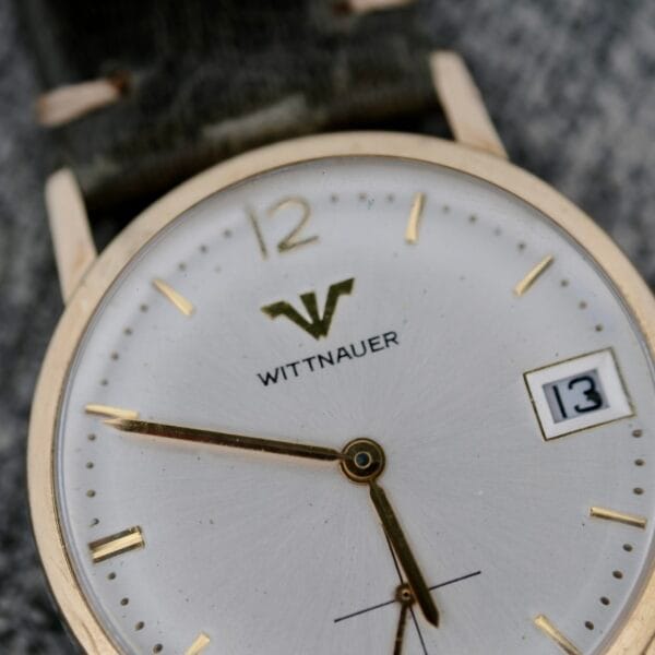 A close-up of the Wittnauer Date Manual Wind 2732-1-C189 wristwatch, featuring a white face, gold hour markers and hands, a date window showing 13, a gold case, and a green strap.