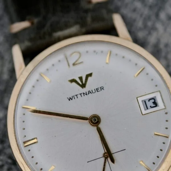 A close-up of the Wittnauer Date Manual Wind 2732-1-C189 wristwatch, featuring a white face, gold hour markers and hands, a date window showing 13, a gold case, and a green strap.