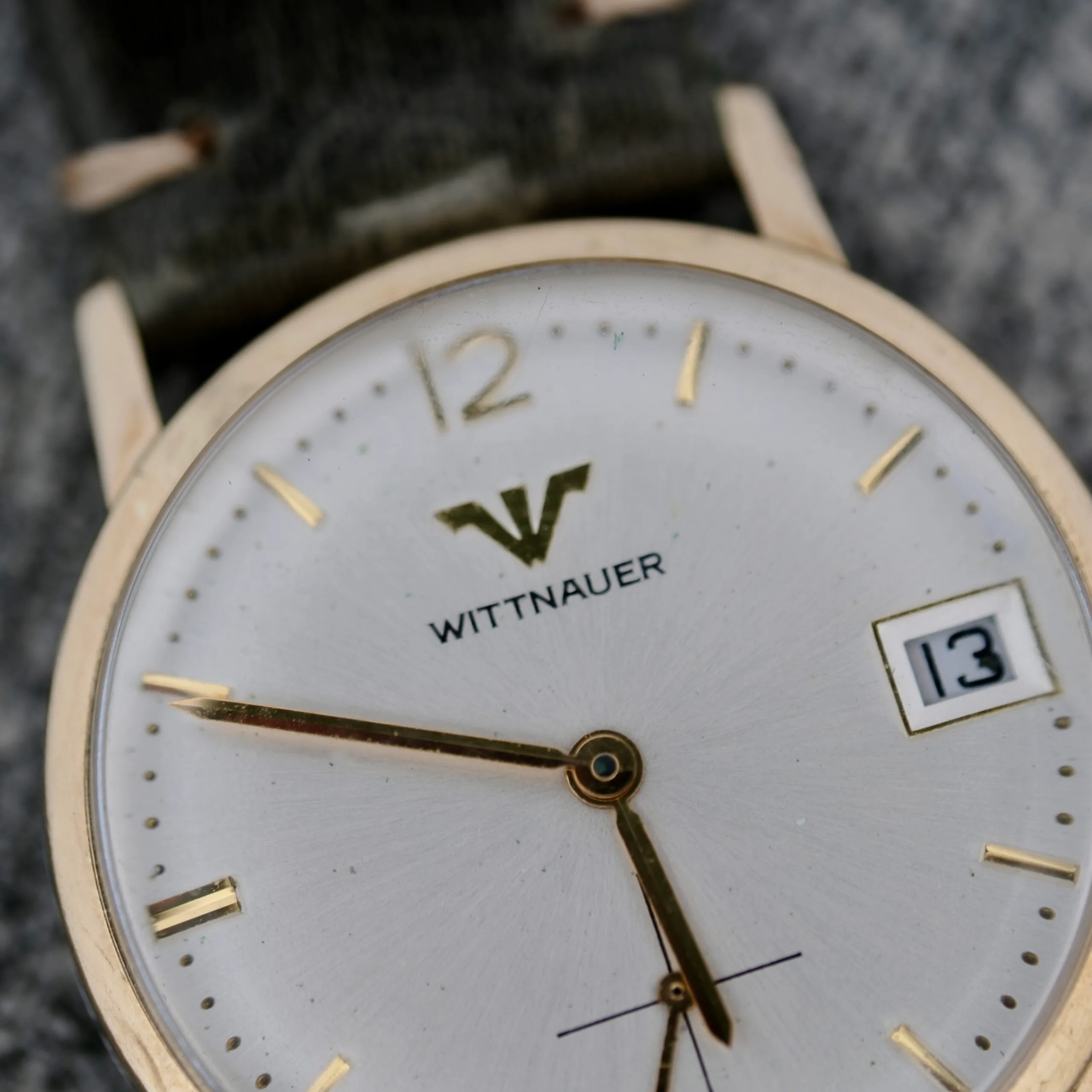 A close-up of the Wittnauer Date Manual Wind 2732-1-C189 wristwatch, featuring a white face, gold hour markers and hands, a date window showing 13, a gold case, and a green strap.