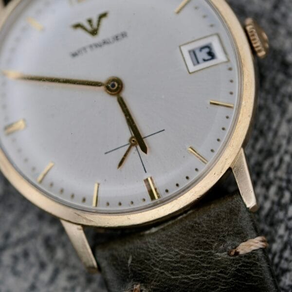 A close-up of the Wittnauer Date Manual Wind 2732-1-C189 wristwatch shows a white and gold dial, gold markers, date display at 13, black leather strap, and the time reading about 6:01:27.