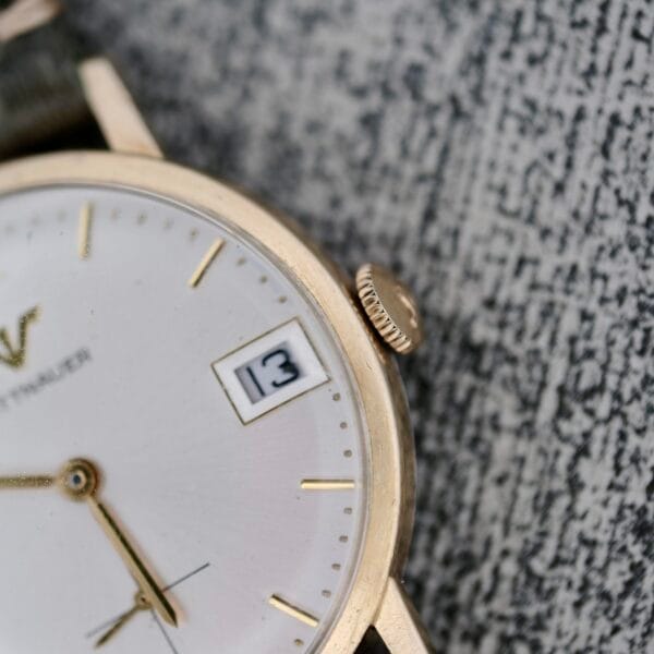 A close-up of the Wittnauer Date Manual Wind 2732-1-C189 gold wristwatch shows the time at 4:33 and the date as 13 on a white face with gold markers, resting on a textured grey surface.
