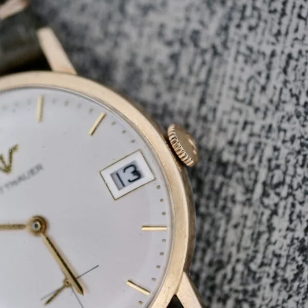 A close-up of the Wittnauer Date Manual Wind 2732-1-C189 gold wristwatch shows the time at 4:33 and the date as 13 on a white face with gold markers, resting on a textured grey surface.