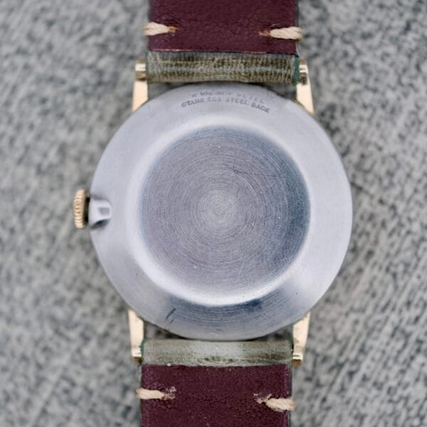 A close-up of the Wittnauer Date Manual Wind 2732-1-C189’s round stainless steel back, brown leather strap with visible stitching, resting on a textured gray surface. Faint engraved text appears near the top edge of the case back.