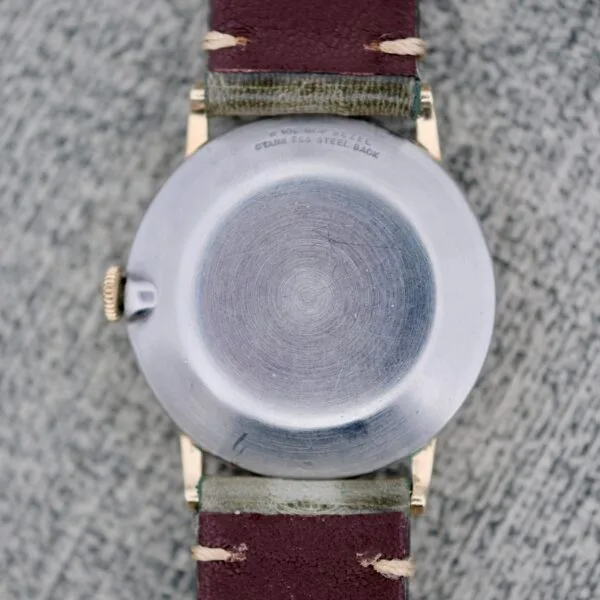 A close-up of the Wittnauer Date Manual Wind 2732-1-C189’s round stainless steel back, brown leather strap with visible stitching, resting on a textured gray surface. Faint engraved text appears near the top edge of the case back.
