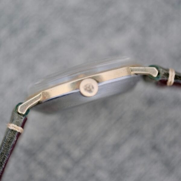 Close-up side view of the Wittnauer Date Manual Wind 2732-1-C189 wristwatch with a round, slim metal case, visible crown and lugs, paired with a stitched leather strap. The background is blurred.